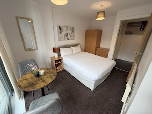 Ein Hotelzimmer mit Bett, Schreibtisch, Stuhl, Nachttisch, Lampe, Kleiderschrank und Tür.