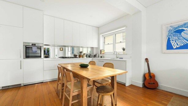 A modern kitchen with white cabinetry, a wooden dining table, and a guitar on the wall. - Large Two-Bedroom Garden Apartment (North Bondi)