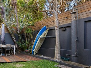 Une terrasse en bois avec une planche de surf appuyée contre un mur, le tout entouré de verdure.