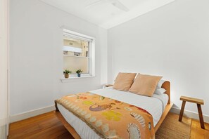 A bedroom with a bed, a window, a small shelf, and a wooden stool. - Private Garden Apartment near Bondi Beach (North Bondi)