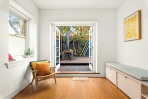 A room with a wooden floor, a bench, a chair, a window, and a door leading to an outdoor area with a table and chairs. - Private Garden Apartment near Bondi Beach (North Bondi)