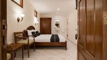 A hotel room with a wooden bed, a bedside table, a chair, a nightstand, a wall-mounted lamp, a door, and a tiled floor.