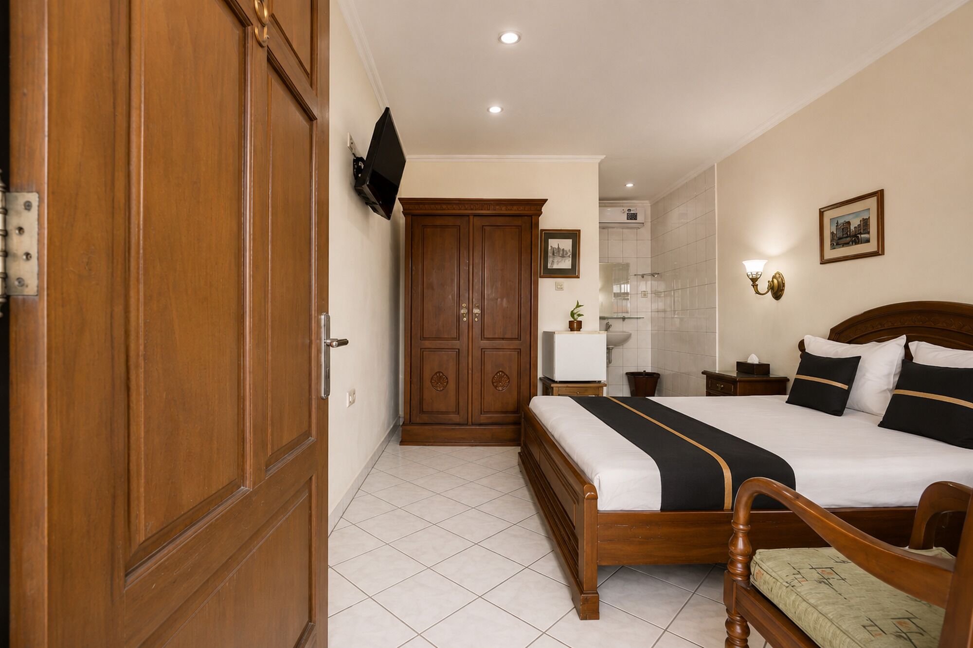 A hotel room with a wooden wardrobe, a bed with a black and white bedspread, a nightstand, a wall-mounted TV, and a bathroom with a sink and mirror.