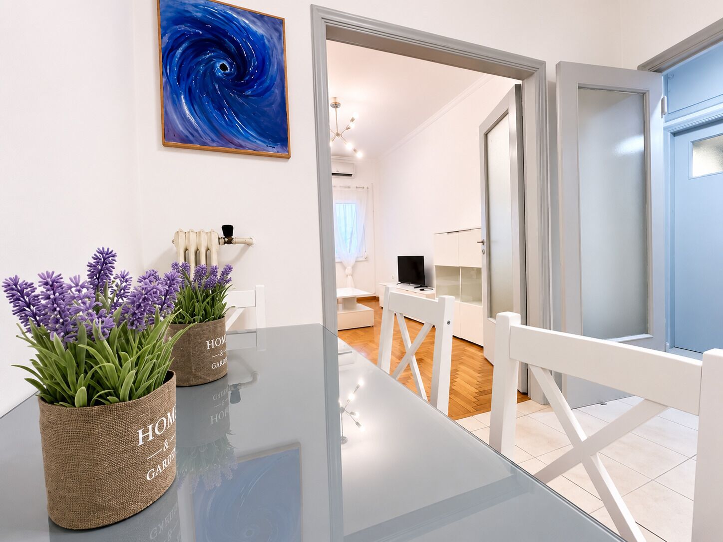 A modern living room with a glass table, two potted plants, a painting, and a view into another room with a TV and white furniture.