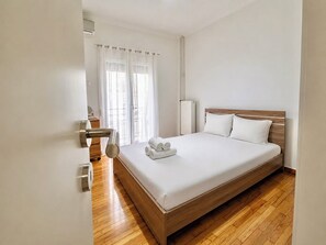 A hotel room with a wooden bed, white linens, a wooden floor, a radiator, and a window with sheer curtains. - Athens Metro Escape - Central & Connected by ΜΡS (Athina)