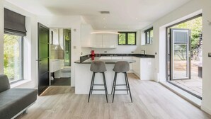 A modern kitchen with a central island, two bar stools, and a view of the outdoors. - Stargazing copper tub backing onto RSPB reserve (Cambridge)