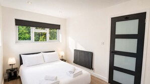 A hotel room with a bed, two bedside lamps, a window with a view of greenery, a radiator, and a black door with frosted glass panels. - Stargazing copper tub backing onto RSPB reserve (Cambridge)