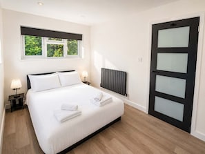 Habitación de hotel con cama, dos lámparas de noche, una ventana con vistas a zonas verdes, un radiador y una puerta negra con paneles de vidrio esmerilado.