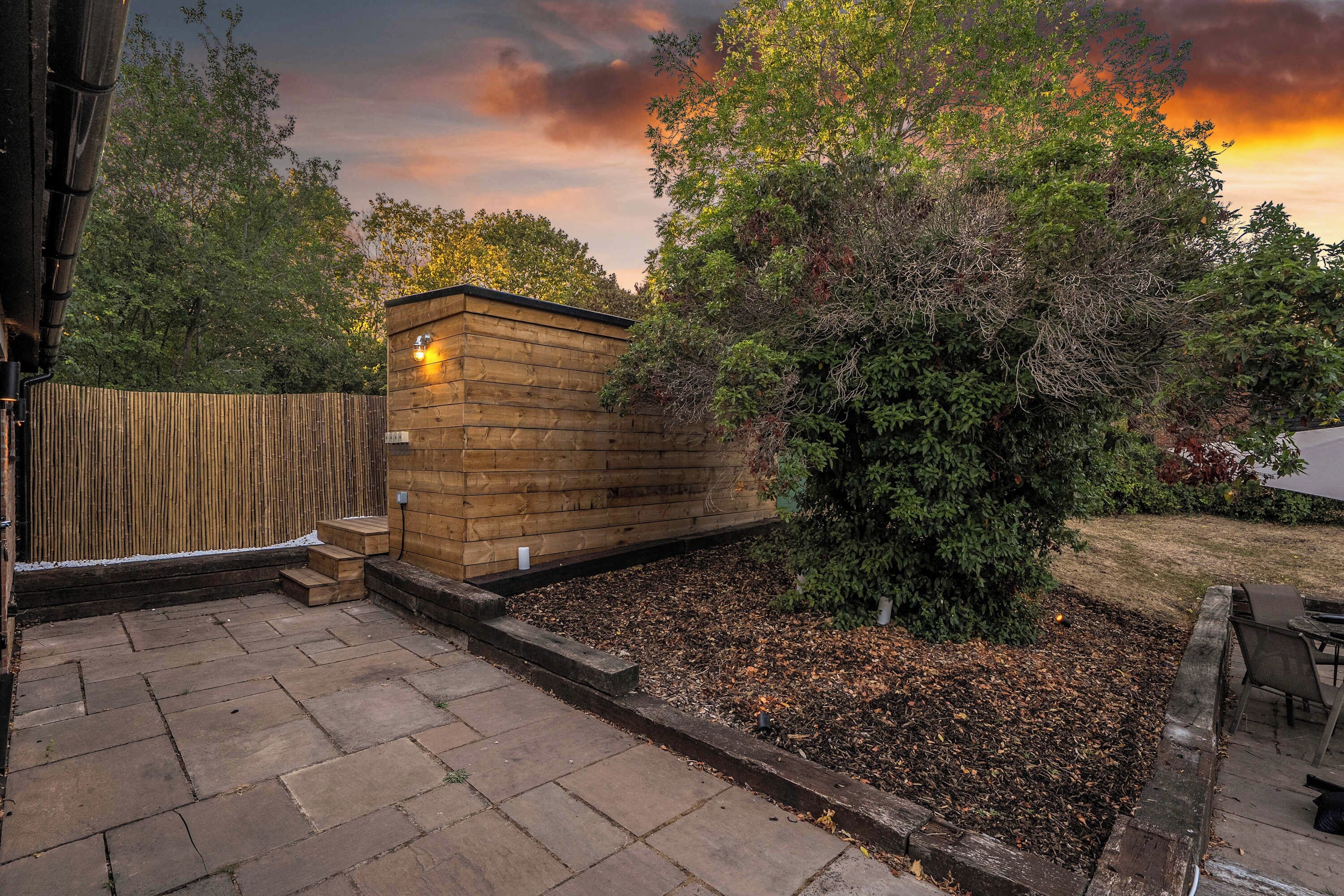 A wooden structure with a light fixture, surrounded by a garden with a tree and a wooden fence.