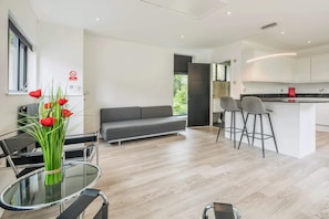 A modern living space with a sofa, a dining table with chairs, a kitchen area, and a glass table with a vase of red flowers.