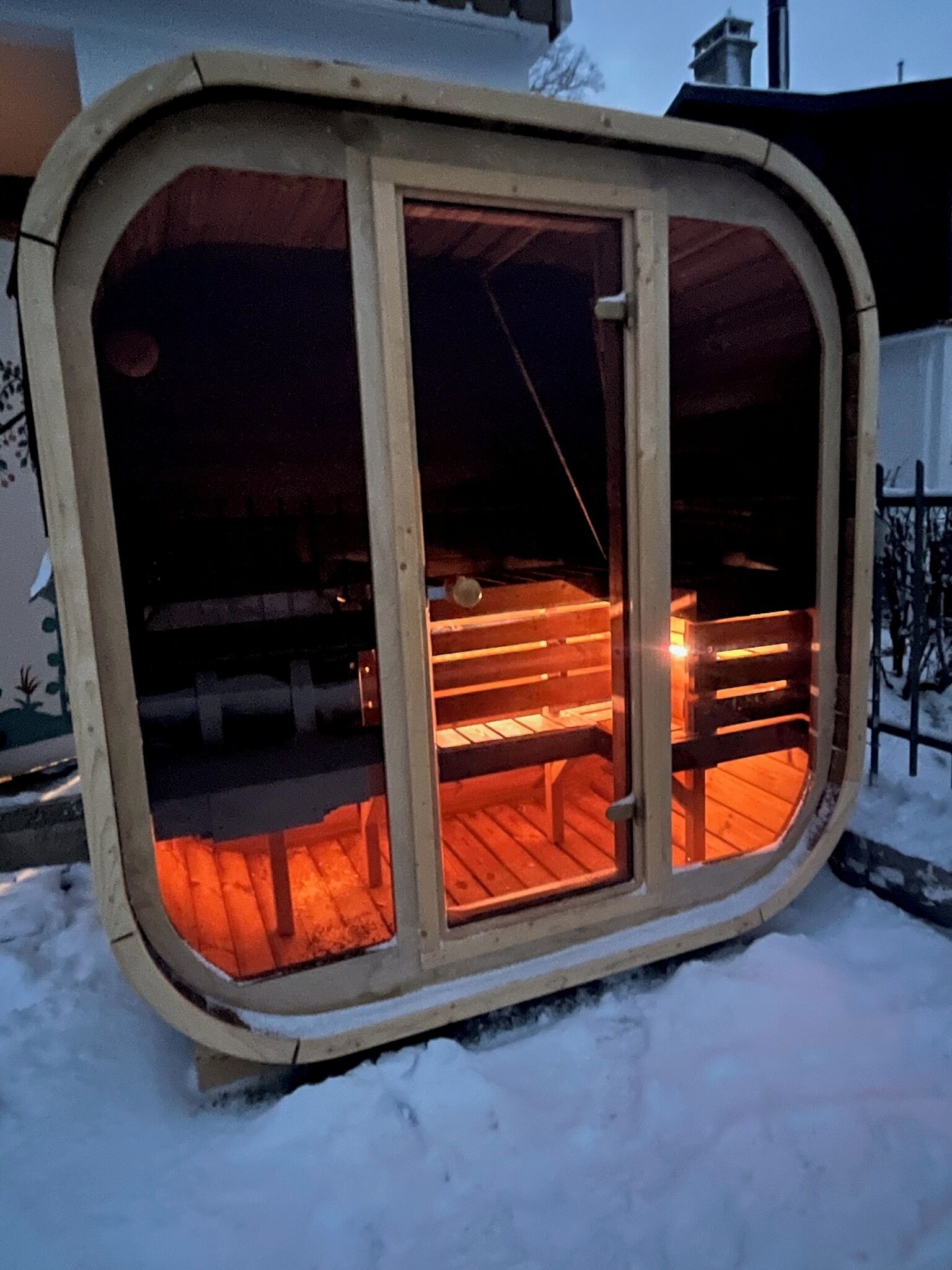 Un espacio exterior cubierto de nieve con una estructura de madera que tiene una puerta de cristal y un banco en su interior.
