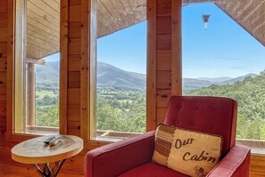 A cabin interior with a red upholstered chair, a small round table with a remote control, and a view of a mountainous landscape through large windows. - ALTA VISTA – Incredible Smoky Mountain & Wears Valley Views with Easy Access (Sevierville)