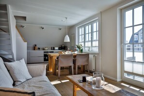 A modern living room with a sofa, a dining table with chairs, and a kitchen in the background. - Experience the ultimate vacation at Haus am Meer Strandburg on Sylt (Wenningstedt-Braderup (Sylt))