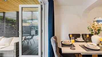 A dining area with a table set for two, a wicker sofa, and a view through a glass door to an outdoor deck.