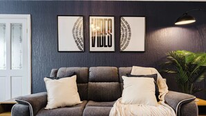 A modern living room with a grey sofa, white cushions, and a knitted throw. There are three framed artworks on the wall, a potted plant, and a table with glasses and bread. - Etihad & Co-op Live, 3-Bed, Free Parking (Droylsden)