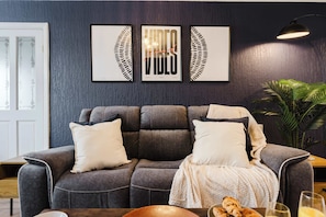 Ein modernes Wohnzimmer mit einem grauen Sofa, weißen Kissen und einer gestrickten Decke. An der Wand hängen drei gerahmte Kunstwerke, eine Topfpflanze und ein Tisch mit Gläsern und Brot.