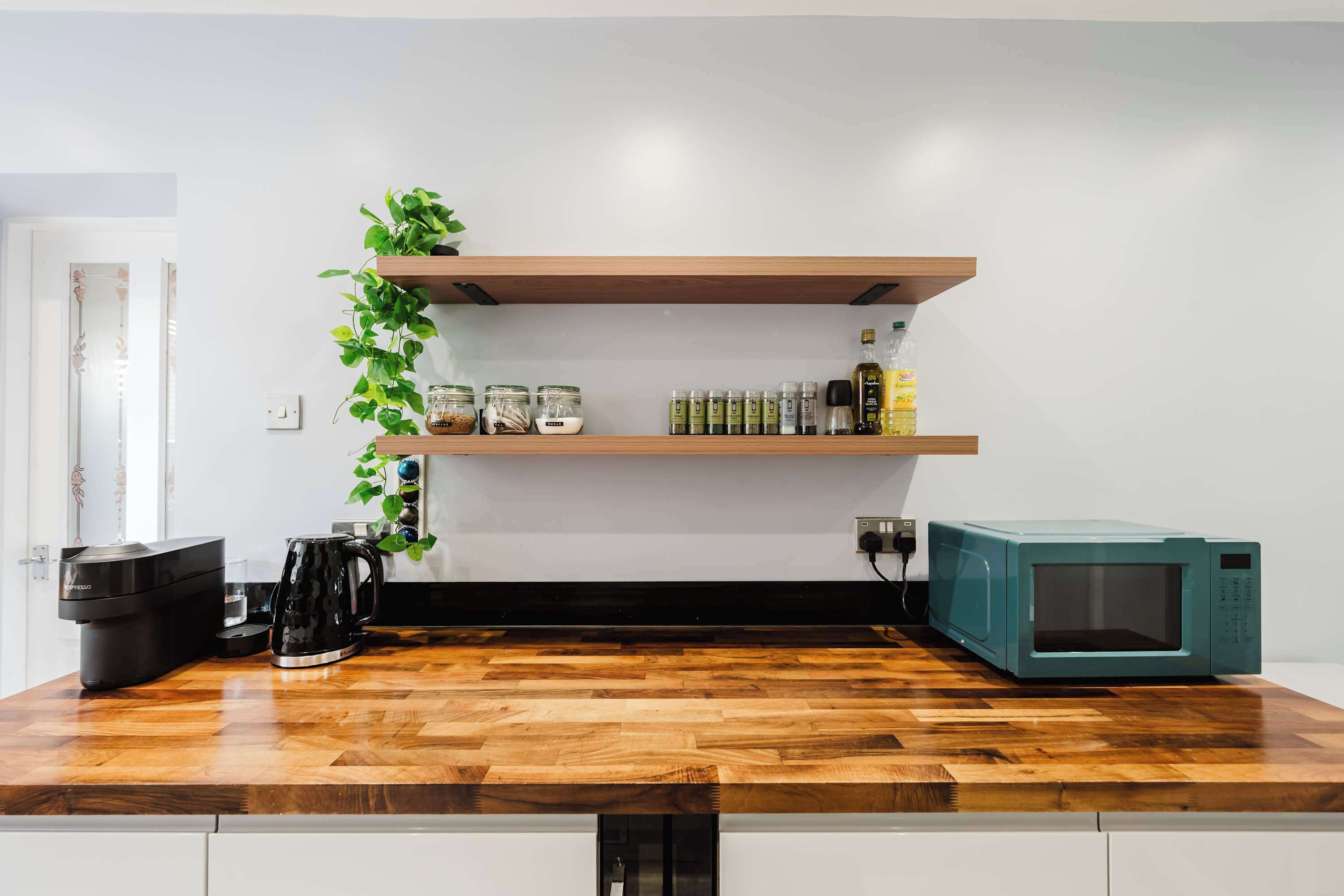 A kitchen with a wooden countertop, a coffee maker, a microwave, and shelves with jars and a plant.