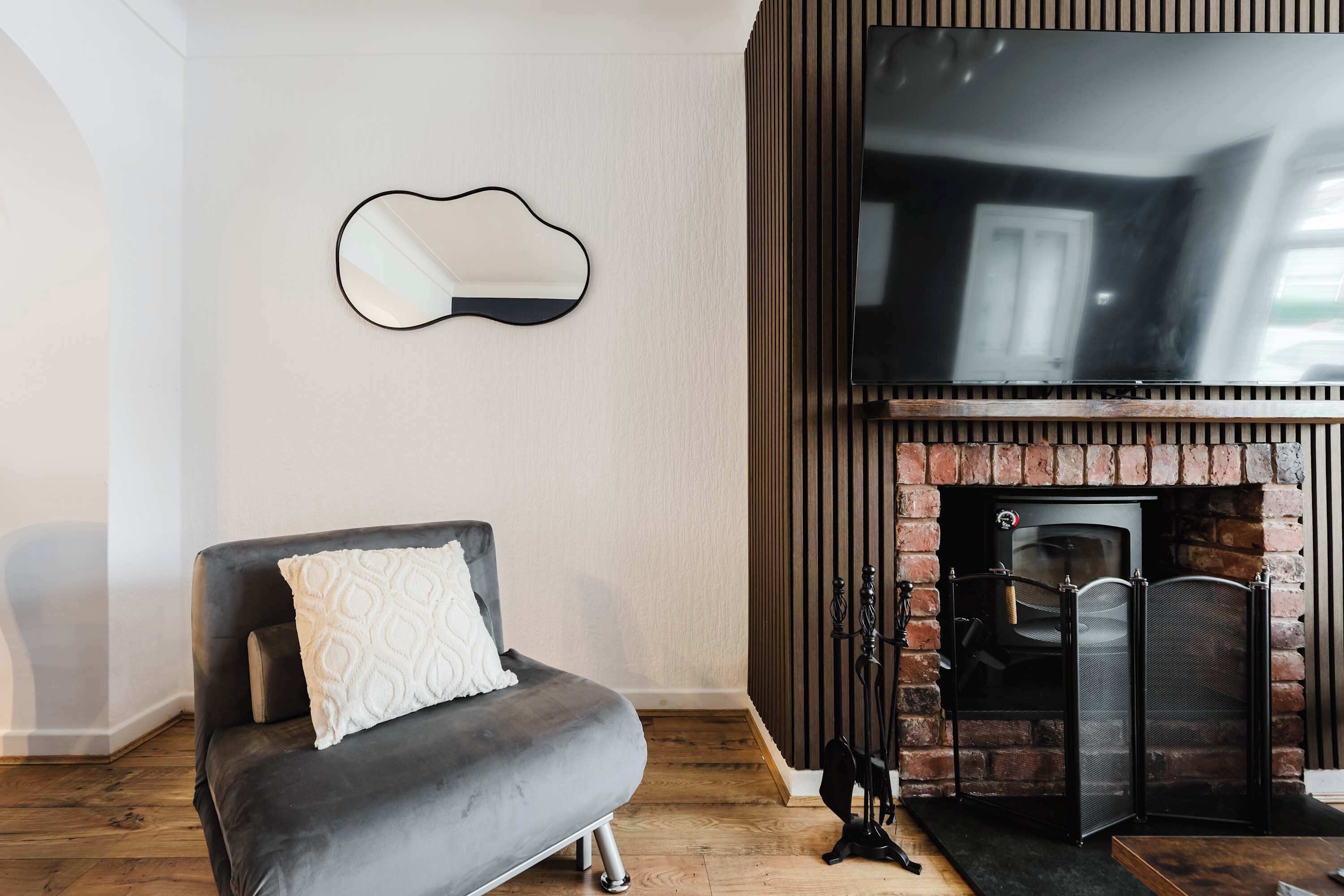 Ein Wohnzimmer mit grauem Sofa, einem flachbildfernseher über einem Kamin aus Ziegelsteinen und einem an der Wand befestigten Spiegel.