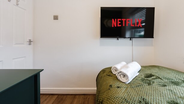 A room with a television mounted on the wall displaying the Netflix logo, a green upholstered bed with a rolled white towel, and a dark-colored dresser. - Etihad & Co-op Live, 3-Bed, Free Parking (Droylsden)