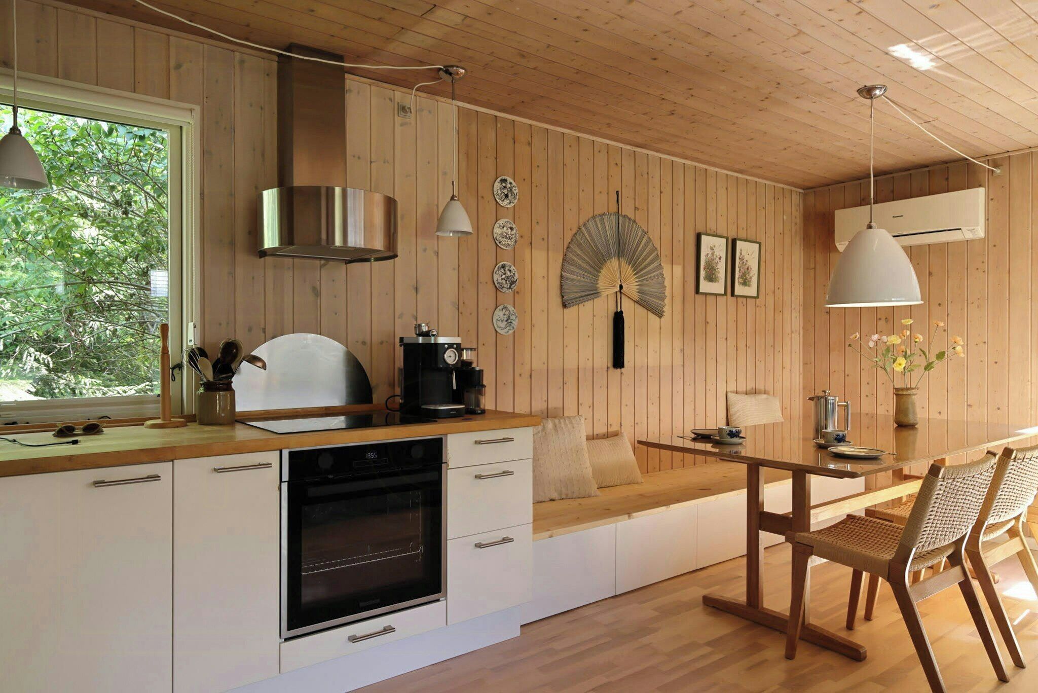Une cuisine moderne avec des murs en bois, un four encastré, un coin repas avec une table et des chaises, et une grande fenêtre qui laisse entrer la lumière naturelle.