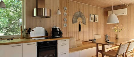 A modern kitchen with wooden walls, a built-in oven, a dining area with a table and chairs, and a large window providing natural light.