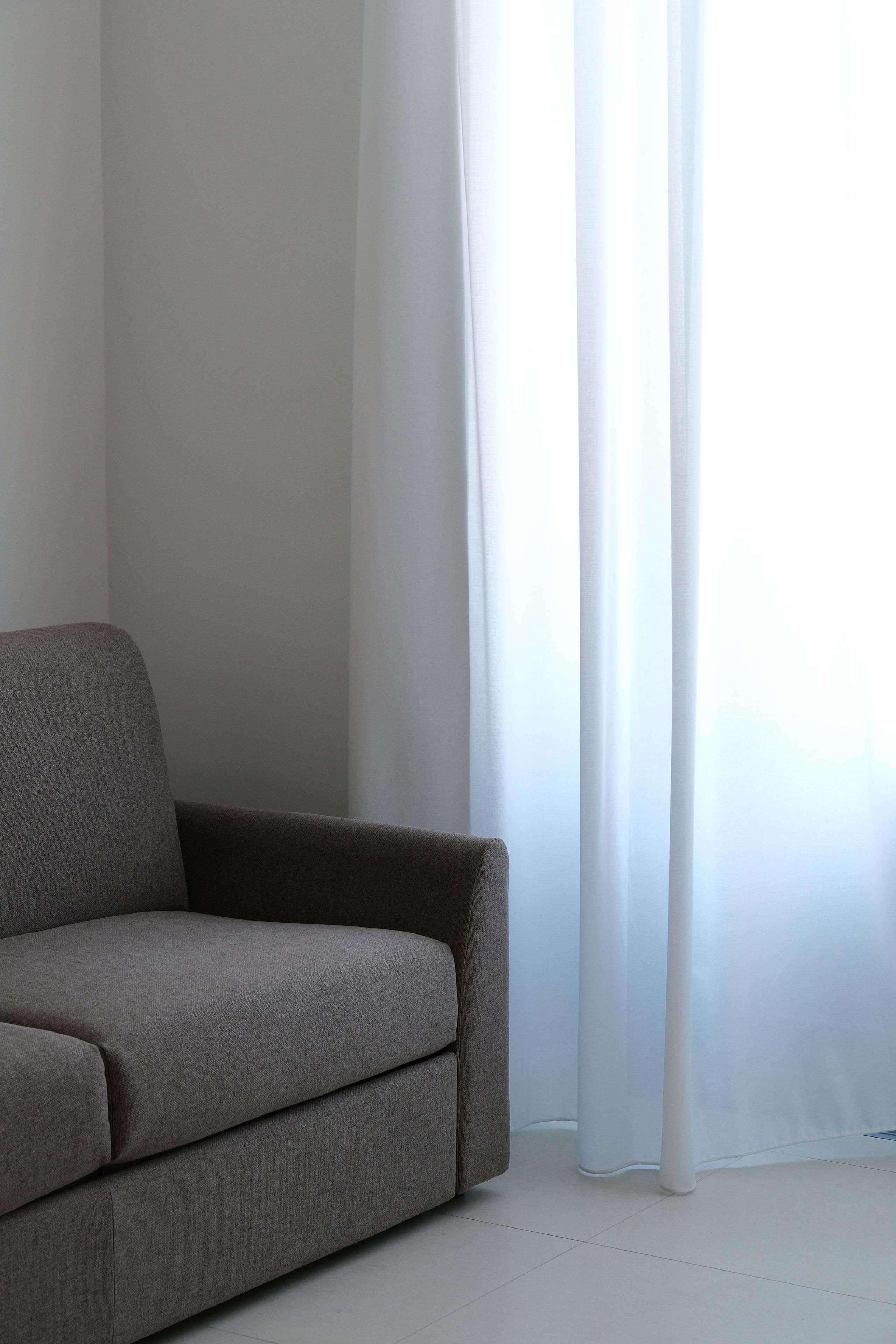 Ein graues Sofa in einem Raum mit weißen Wänden und einem Fenster mit durchscheinenden Vorhängen.