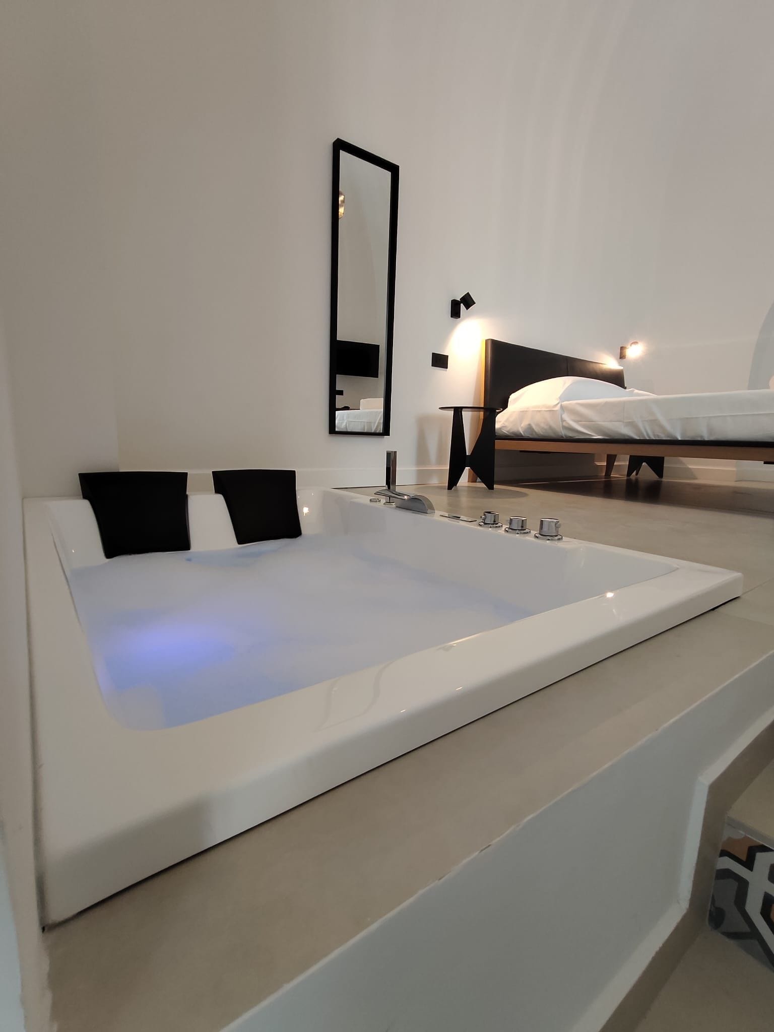 A modern bathroom with a large bathtub, a mirror, and a bed in the background.