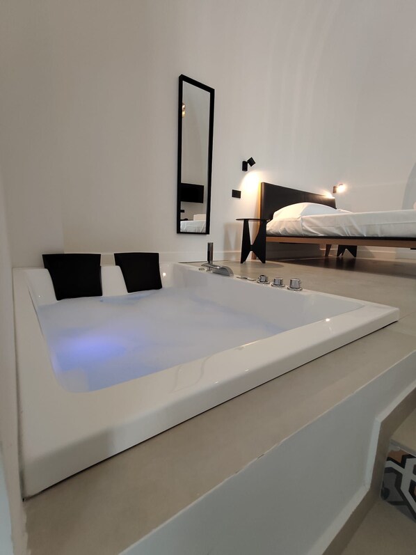 A modern bathroom with a large bathtub, a mirror, and a bed in the background.