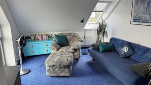 Ein Wohnzimmer mit blauem Teppich, einem gemusterten Sofa, einem blauen Schlafsofa, einer Stehlampe und einem Bücherregal.