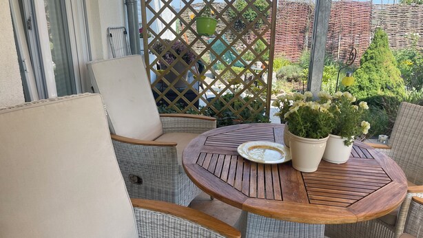 A balcony with wicker furniture, a round wooden table, and potted plants. - Studio Jardin Harmonie, sehr elegant wohnen Pantry-Küche, Terrasse, Garten, Pool (Rain)