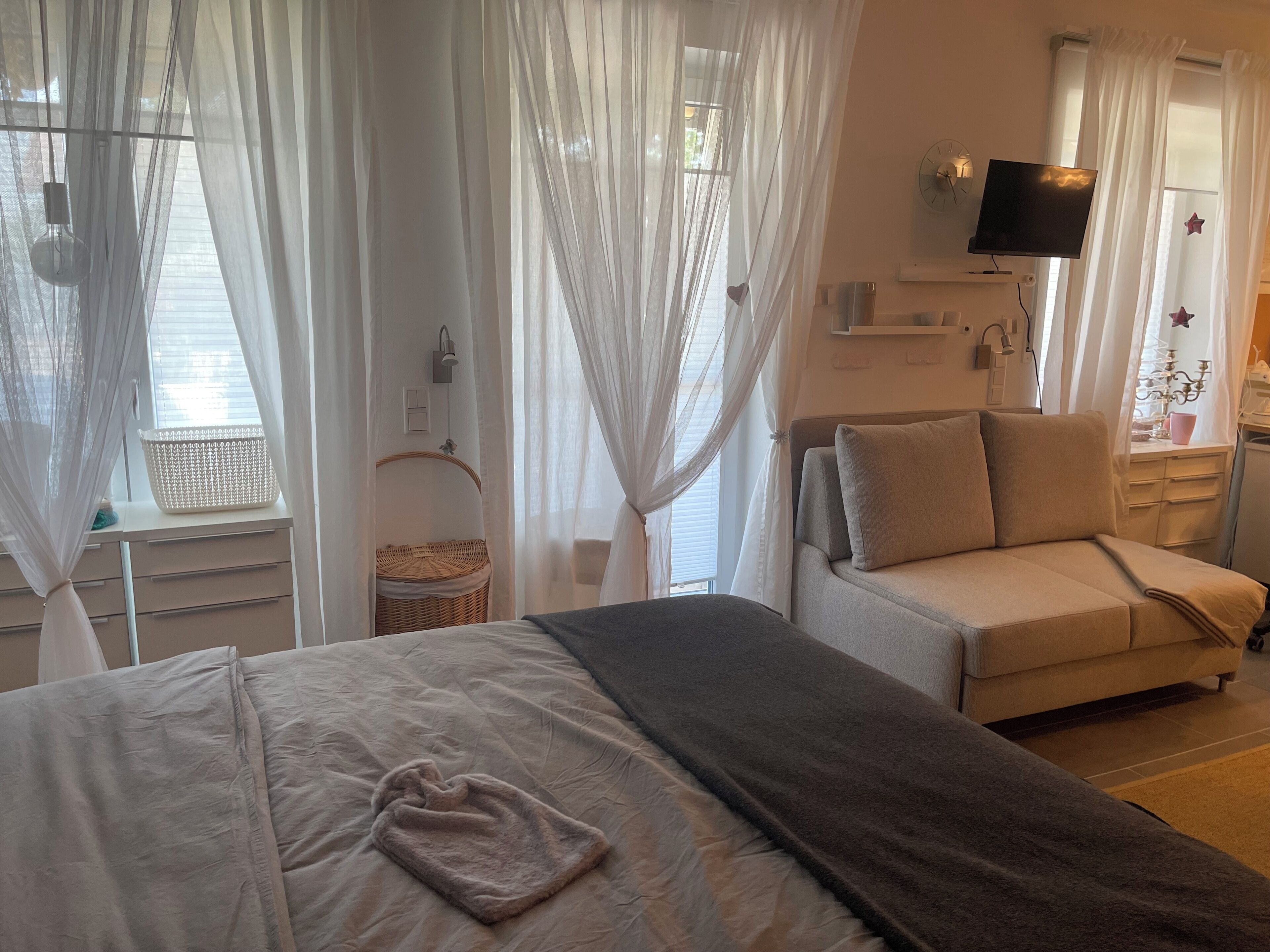 Een slaapkamer met een bed, een bank, een televisie en een raam met dunne gordijnen.