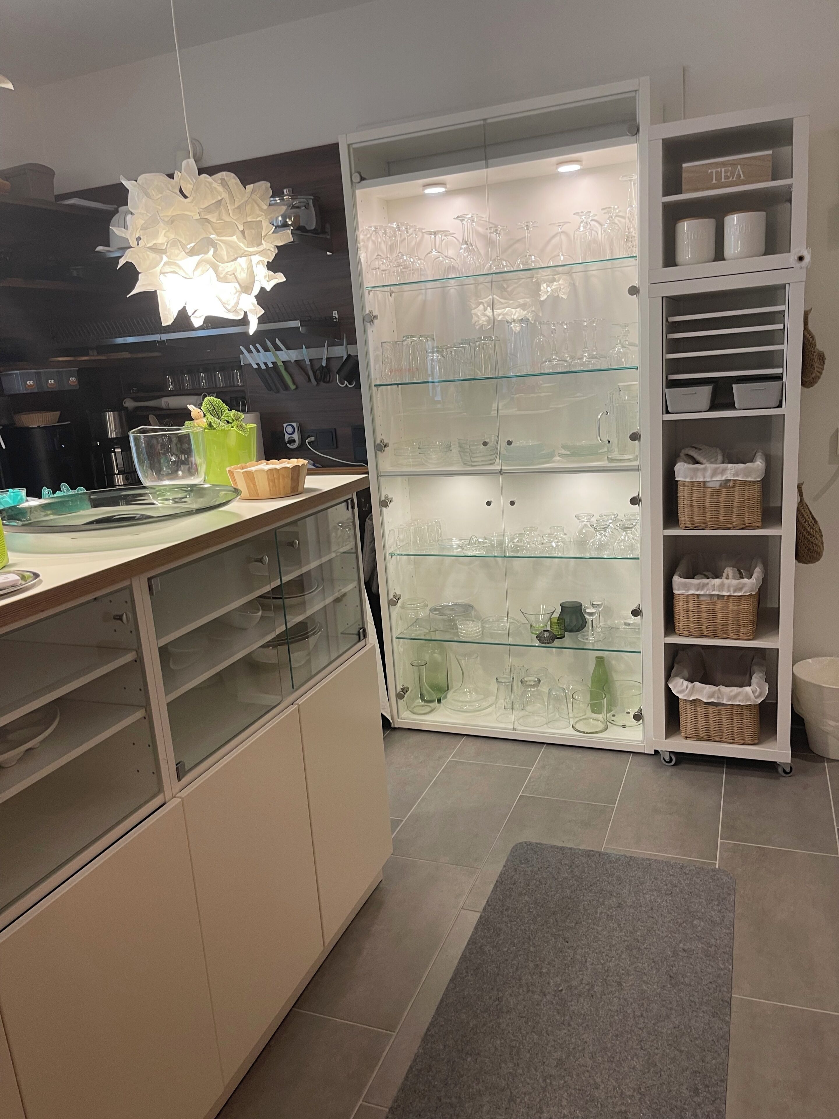 A modern kitchen with a glass display cabinet filled with glassware and a white cabinet with storage baskets.