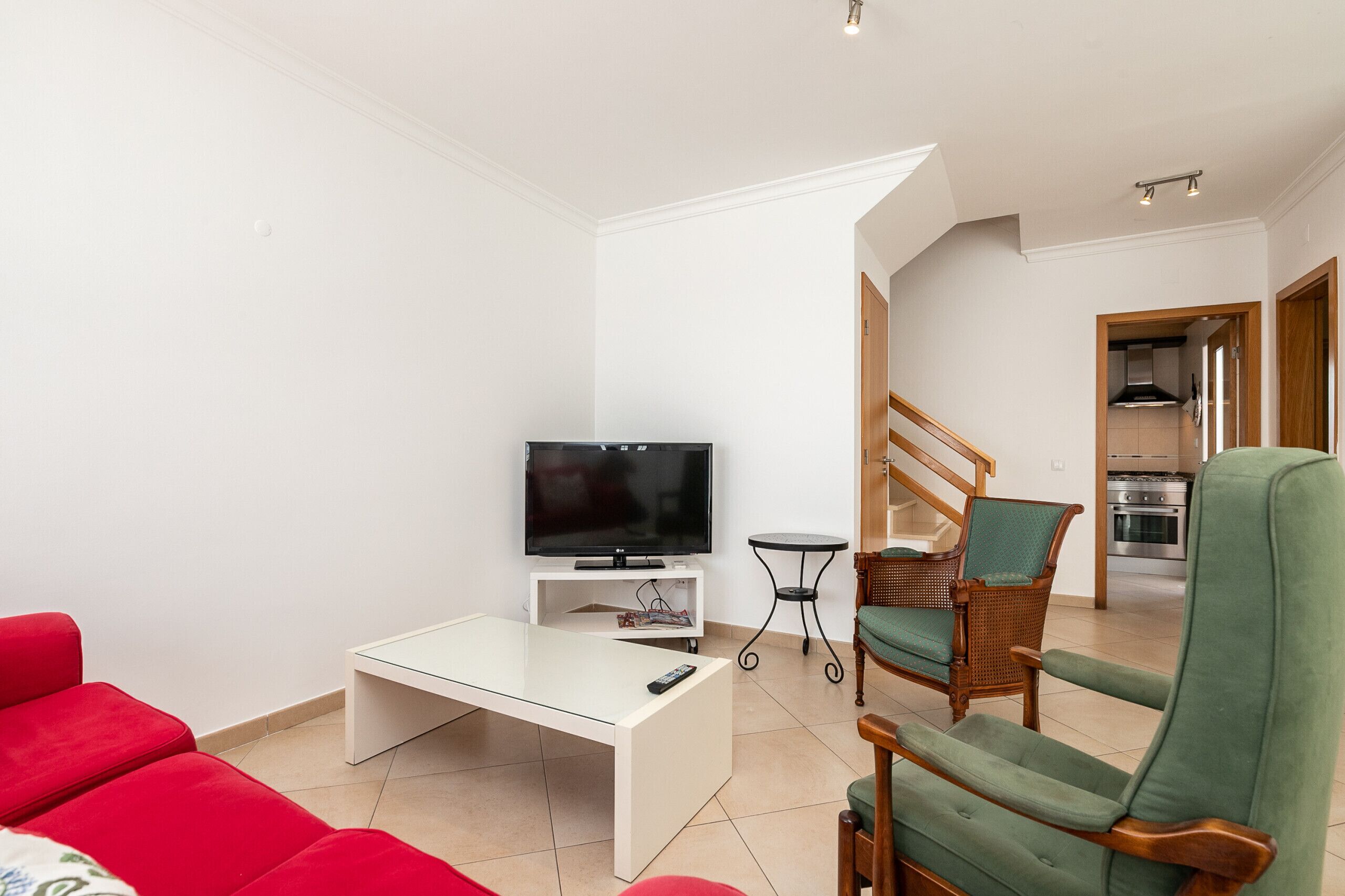 Een woonkamer met een flatscreen televisie op een witte standaard, een rode bank, een groene fauteuil en een kleine tafel.