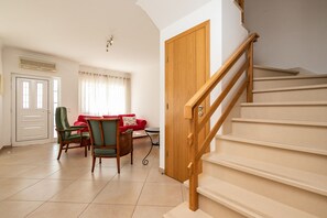 Una scala con ringhiere in legno conduce a un pianerottolo, dove si trovano un divano rosso e una poltrona verde in un soggiorno con pavimento piastrellato.