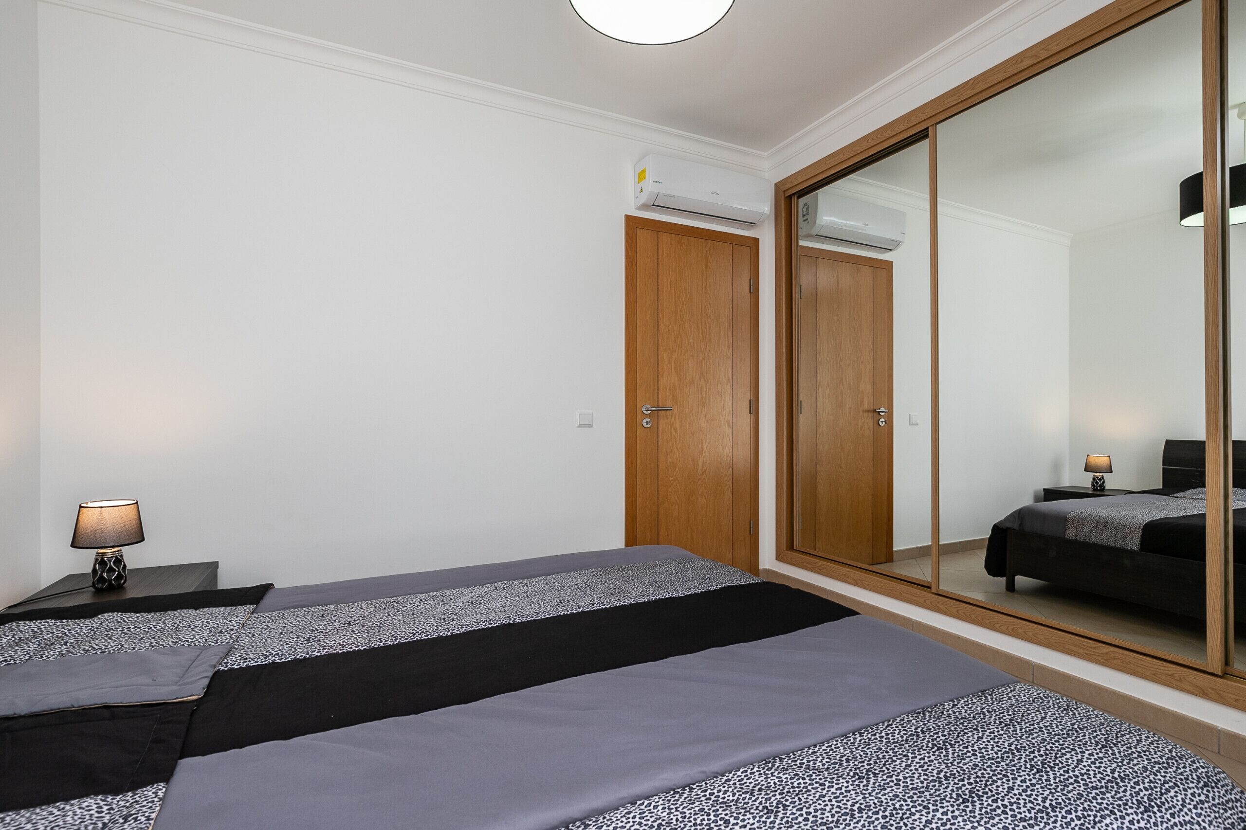 A bedroom with a bed, a nightstand, a lamp, a wardrobe, and a large mirror.