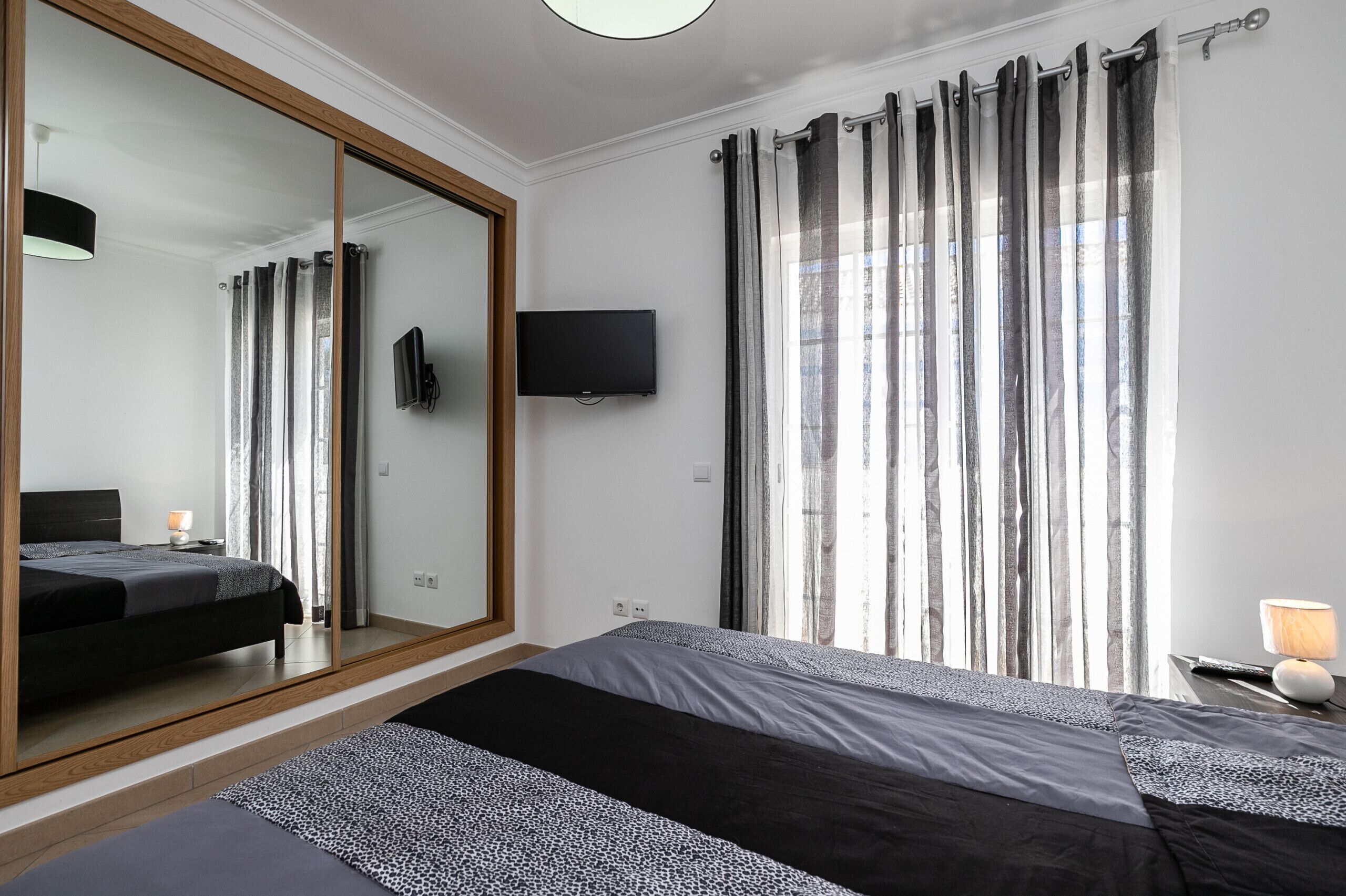 A bedroom with a large mirror, a bed, a TV, and a window with curtains.