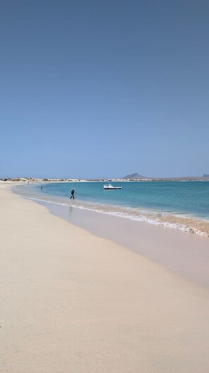 A beach with a boat in the water, a person walking, and a distant mountain. - Our eco friendly home 2 min from Cabral Beach (Sal Rei)