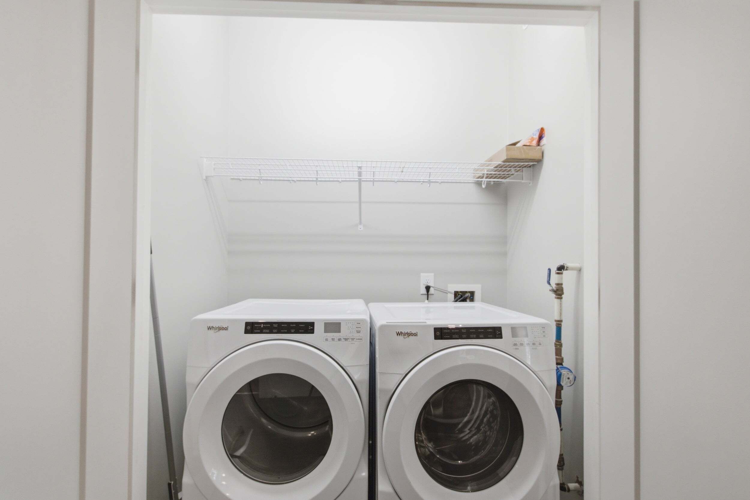 A laundry room with two front-loading Whirlpool washers and a metal shelf above them.
