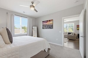 A bedroom with a bed, a ceiling fan, a window with curtains, and a cityscape painting on the wall.