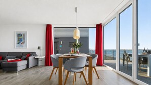 A modern living room with a dining area, large windows, and a view of the sea. - Maritimes & Sea – Vacation Home with Sauna and Ocean View on the Beach (Olpenitz)