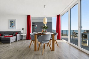 A modern living room with a dining area, large windows, and a view of the sea. - Maritimes & Sea – Vacation Home with Sauna and Ocean View on the Beach (Olpenitz)