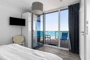 A modern bedroom with a bed, a flat-screen TV, a sofa, and a view of the sea through a sliding glass door. - Maritimes & Sea – Vacation Home with Sauna and Ocean View on the Beach (Olpenitz)