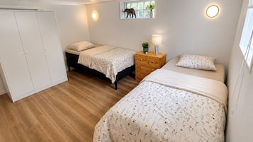 A bedroom with two beds, a wardrobe, a bedside table with a lamp, and a window with a view of trees.