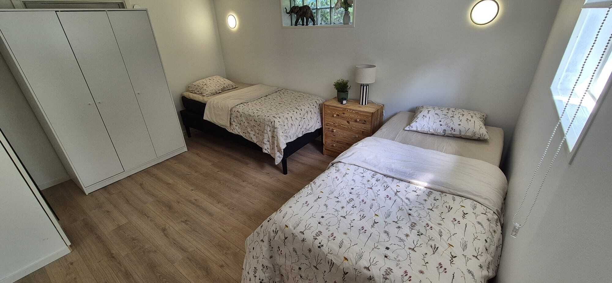 A bedroom with two beds, a wardrobe, a nightstand with a lamp, and a window with a view of trees.