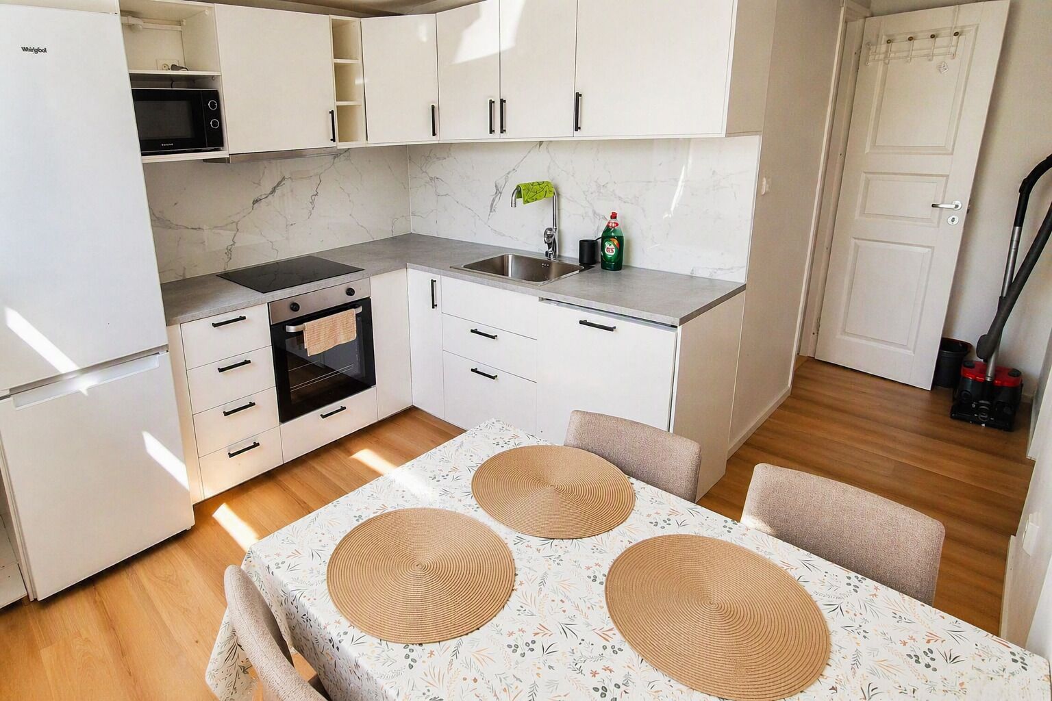 A compact kitchen with white cabinets, a microwave, an oven, and a sink. There is a table with a floral tablecloth and woven placemats.