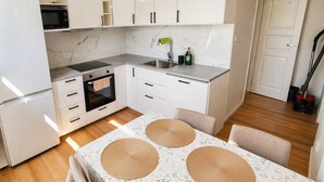 Uma cozinha compacta com armários brancos, micro-ondas, forno e pia. Há uma mesa com toalha floral e jogos americanos trançados.