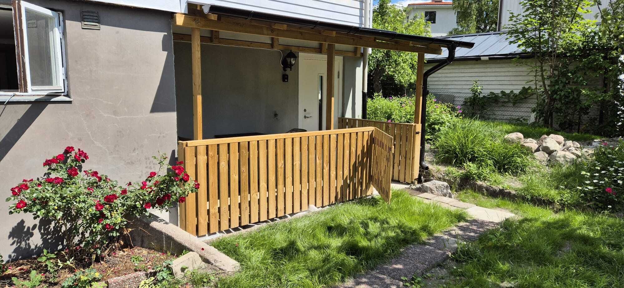 En träveranda som är ansluten till ett hus och har en dörr, omgiven av en trädgård med röda blommor och grönt gräs.