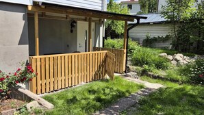 Um deck de madeira anexado a uma casa com uma porta, rodeado por um jardim com flores vermelhas e grama verde.