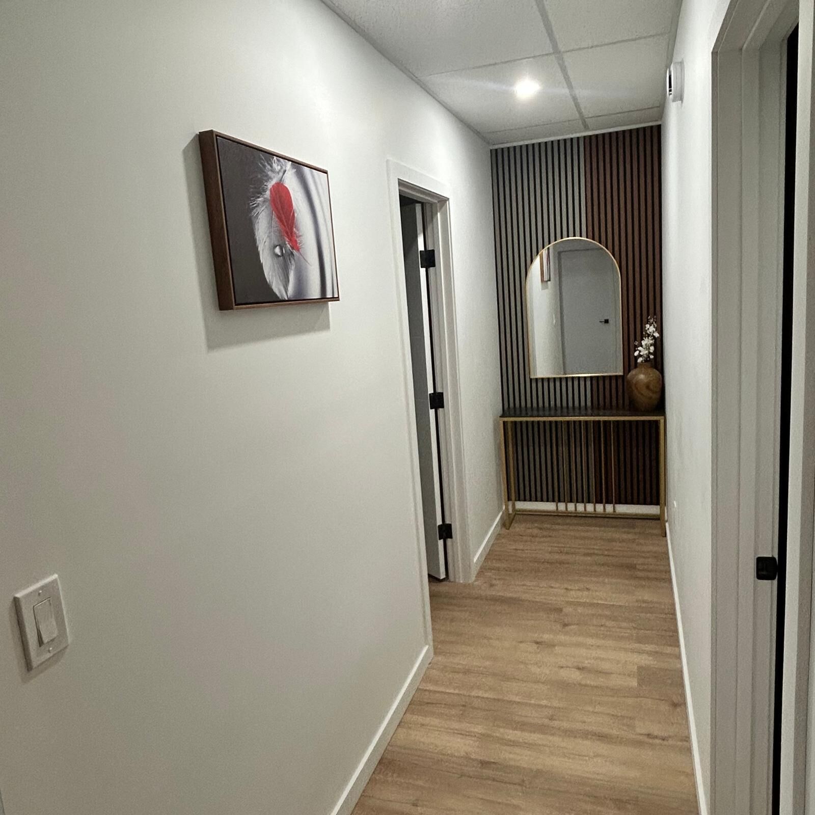 A hallway with a wooden floor, a door, a wall-mounted mirror, and a framed picture.