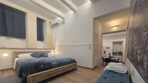 A bedroom with a bed, a bench, a wall-mounted air conditioner, and a view into a kitchen area.